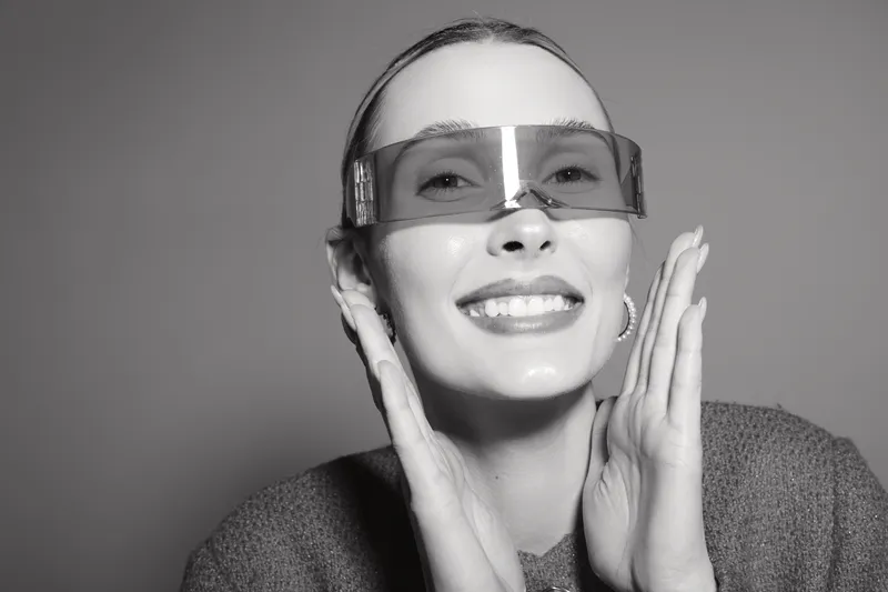 Smiling white woman with light skin wearing transparent sunglasses and cozy sweater touches face. Studio portrait with soft lighting and happy mood concept.