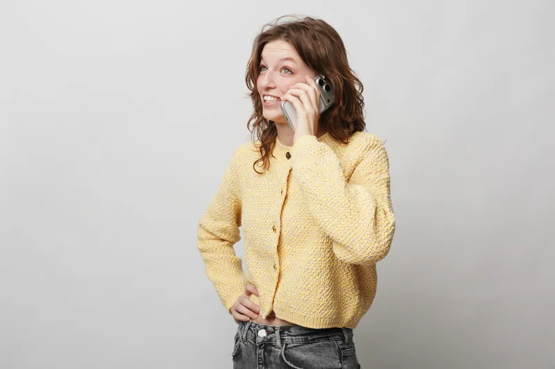 Young woman with curly hair wearing yellow knitted sweater talks on smartphone smiling. Casual style, communication concept, isolated on light grey background with positive mood.