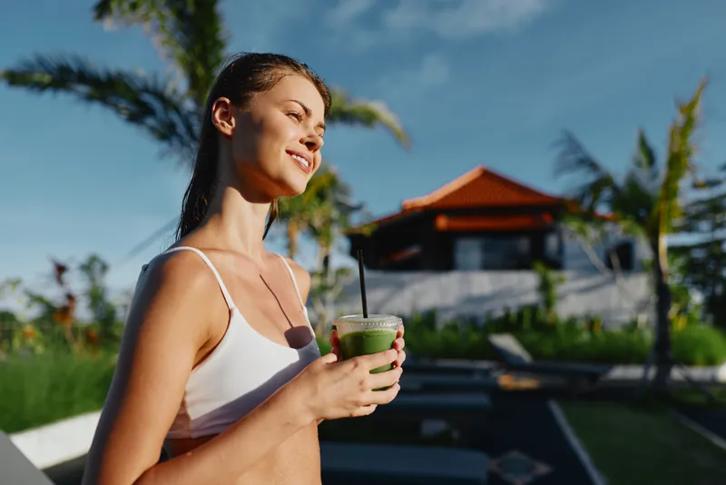 Relaxed woman in a white top enjoys a green smoothie by an outdoor pool at a sunny tropical resort, soaking up leisure and sunshine amid palm trees. A vibrant holiday mood today.