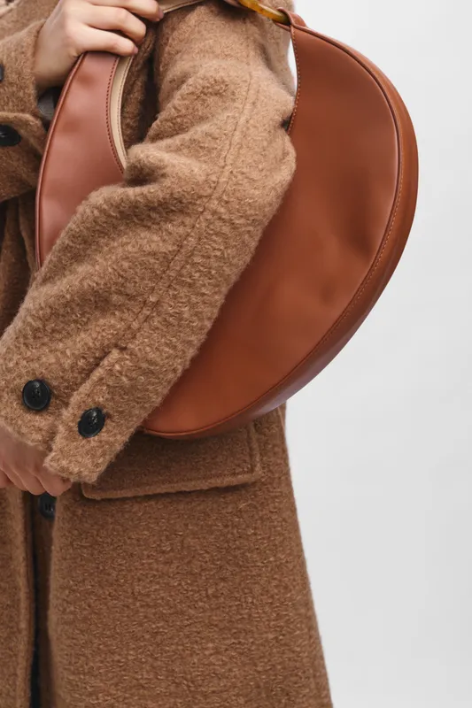 Asian woman wearing a warm brown coat stands with a stylish leather bag, presenting a chic urban look that conveys confidence, elegance, and contemporary fashion in a minimal setting.
