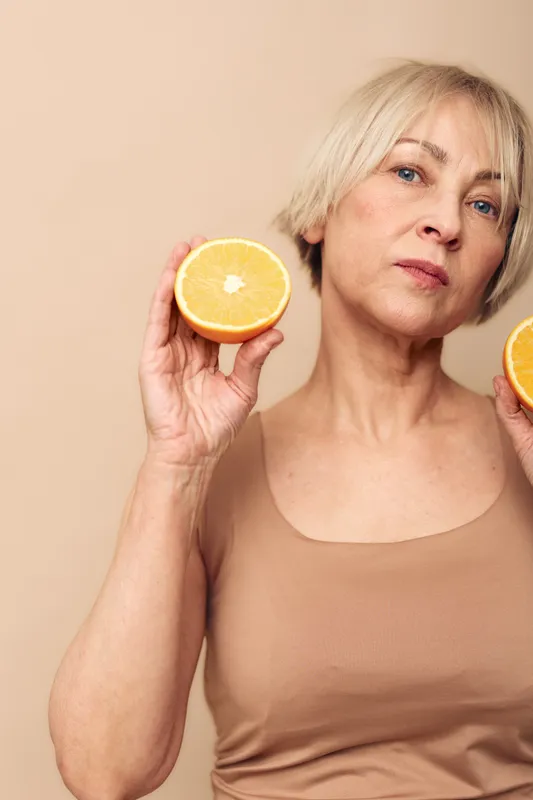 Mature woman with short blonde hair holds two halves of fresh orange fruit near face, wearing beige tank top on beige background. Concept for healthy lifestyle, nutrition, and natural beauty.