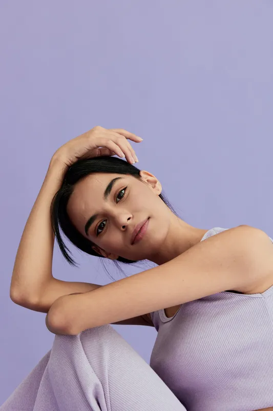 Young woman in lilac outfit poses thoughtfully with arm resting on head against soft purple background, showcasing calm expression and natural beauty.