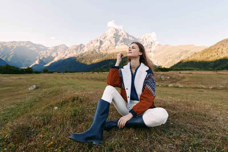 Woman sitting on meadow by mountains, wearing patterned sweater and tall boots, enjoying nature and landscape with relaxed pose in warm autumn light and scenic outdoor view.