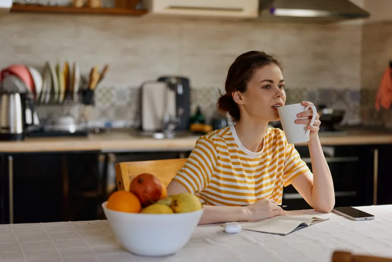 Woman with striped shirt sits at a kitchen table, sipping from a mug beside a bowl of fruit in a cozy home setting. Casual morning routine, wellness, and calm focus.
