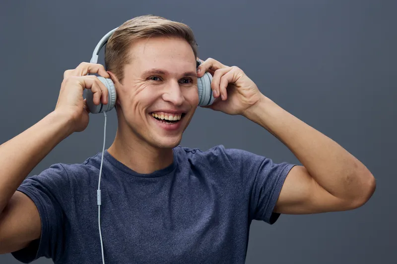 Smiling young man wearing casual dark blue T-shirt enjoying listening to music with large headphones, looking happy, fun, cheerful, isolated on plain solid gray background, studio portrait. People lif