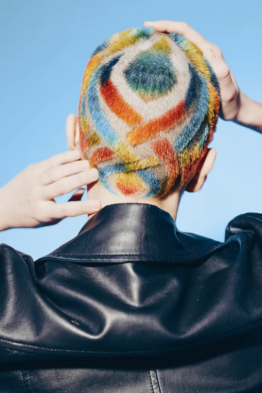 woman with colorful geometric shaved hairstyle wearing black leather jacket stands against clear blue sky, showcasing vibrant fashion and bold self-expression.