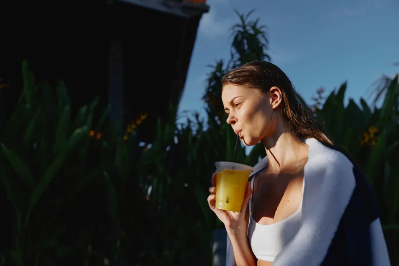 Smiling woman with towel over shoulders holding a glass of juice outdoors, sunlit garden, relaxed morning routine, wellness, self care, casual lifestyle, fresh air, natural lighting.