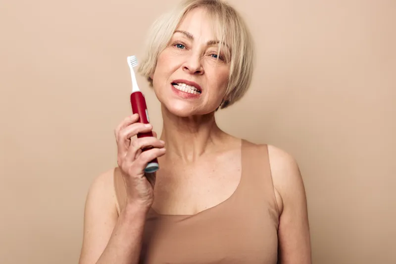 Mature woman with short blonde hair holds electric toothbrush near face, showing teeth in neutral beige background. Concept of dental care, hygiene, oral health, and personal grooming.