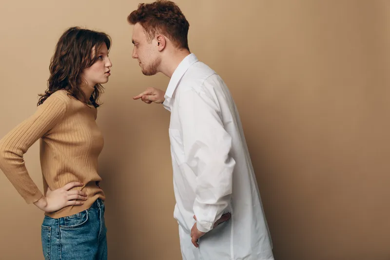 Man in white shirt angrily points finger at woman in brown sweater and jeans. Couple stands face to face in tense argument on beige background. Concept of conflict and disagreement.