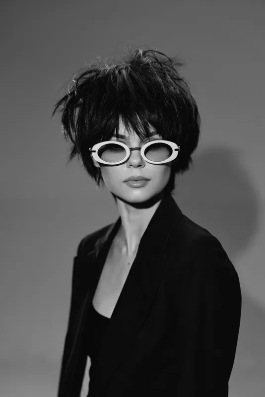 Black and white portrait of a stylish woman wearing oval sunglasses and a blazer with a serious expression and messy hair.