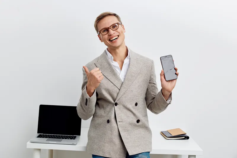 smiling man holding smartphone showing thumbs up wearing grey jacket white shirt standing near laptop on white background remote work digital nomad concept