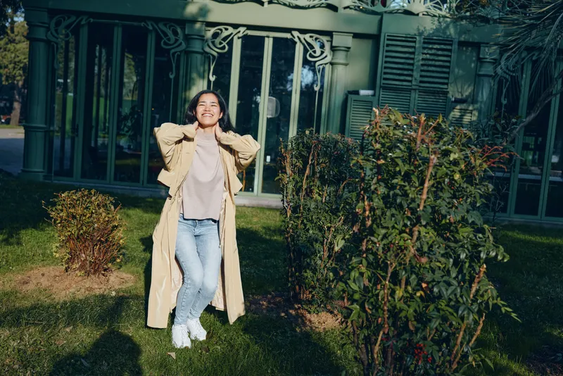 Woman standing in a garden wearing a long beige coat, light sweater and jeans, posing with hands behind head, bright daylight, suburban house backdrop, casual fashion moment.