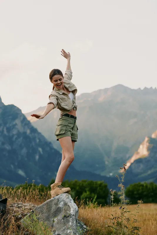 Cheerful woman outdoors, balancing on a rock, hiking style, sunlit landscape, mountains on the horizon, grassy field, carefree moment, natural colors, active lifestyle