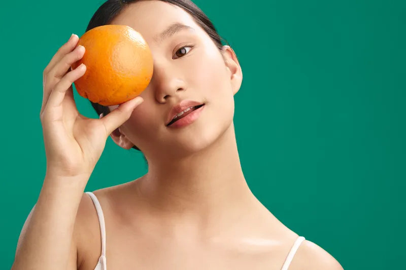 asian woman is shown holding an orange near her face in a studio portrait with a vivid green background, conveying freshness and a playful mood.