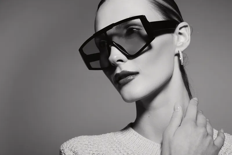 Fashionable white woman with pale skin wearing oversized geometric sunglasses and a textured sweater poses with hand near neck in studio. Concept of style, accessories, and elegance.