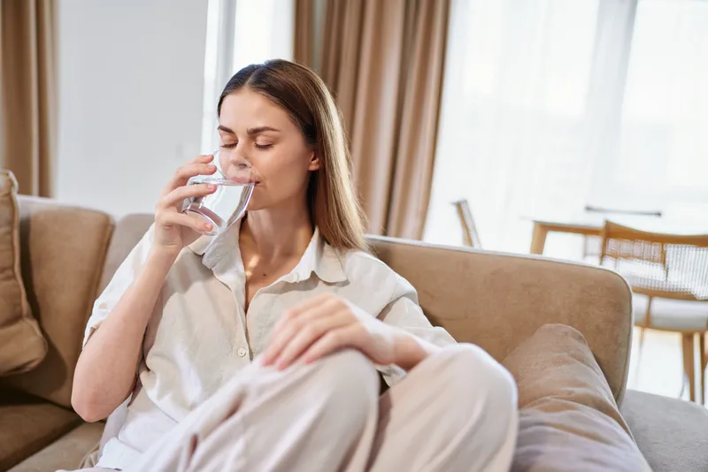 Woman drinking water on sofa at home, relaxed and comfortable, eyes closed while holding a glass on couch in bright living room, cozy lifestyle scene of hydration and calm.