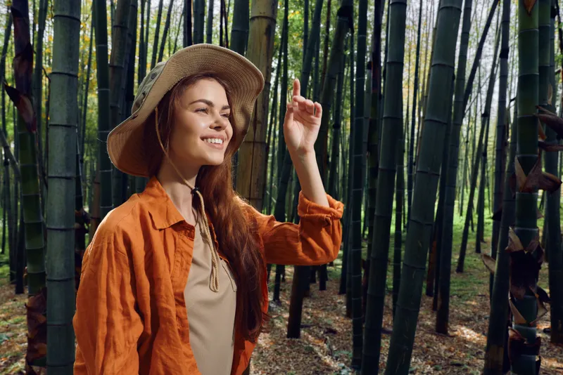 Woman in straw hat smiling among tall bamboo forest, nature travel portrait outdoors. Young female in orange jacket exploring a serene green grove with soft sunlight.
