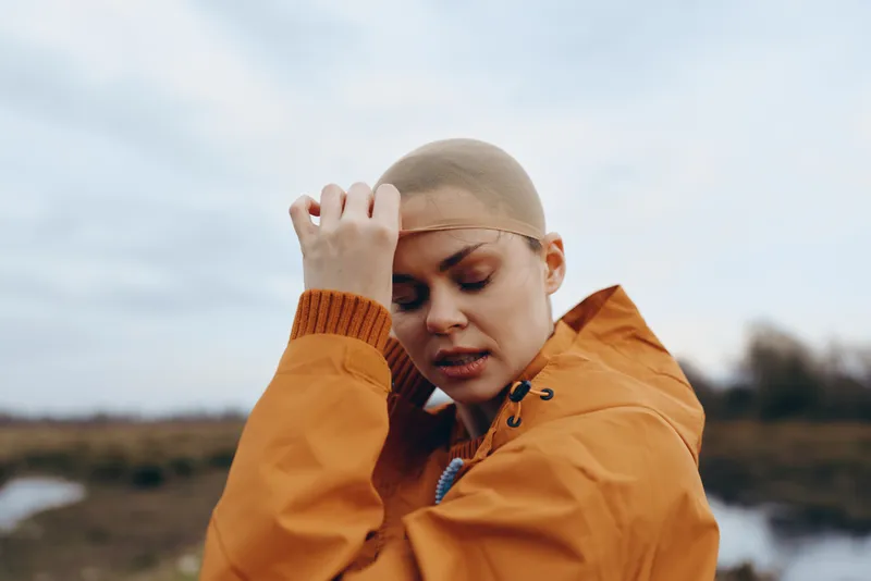 Woman outdoors lifestyle, wearing orange jacket, touching her head with one hand, sadness in her face, tears visible, wind across, nature background, emotional portrait in soft light