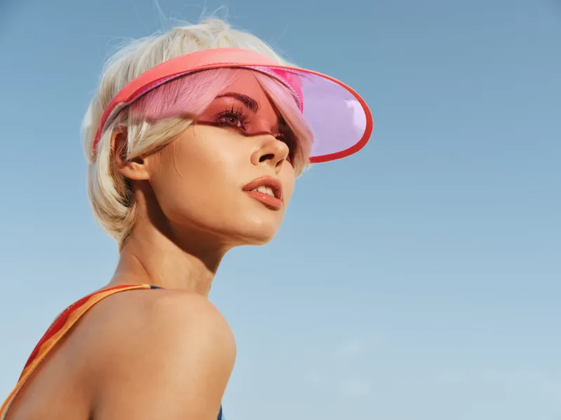 Portrait of a stylish woman wearing a pink visor, looking upward under a clear blue sky; sunlit skin, warm tones, and bold summer fashion with a playful, vibrant vibe.