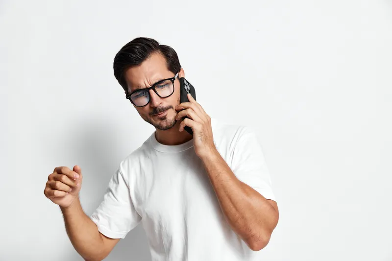 Handsome smiling man talking on mobile phone while expressing emotions, wearing casual white t-shirt, against plain light background, studio portrait. Lifestyle concept.