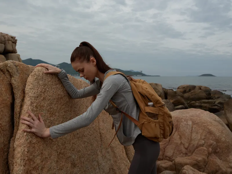 Young woman in athletic wear, exploring rocky coastline with a backpack, expressing determination, overcast skies and natural scenery, outdoor adventure