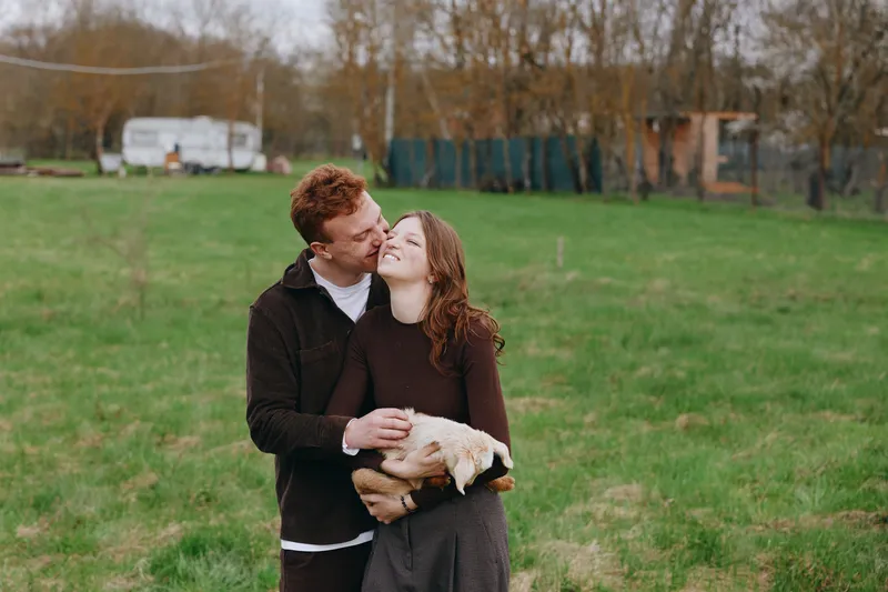 Joyful couple outdoors kissing while holding a playful goat kid in a sunlit field, warm embrace, natural colors, relaxed lifestyle moment, rustic backdrop, candid affection