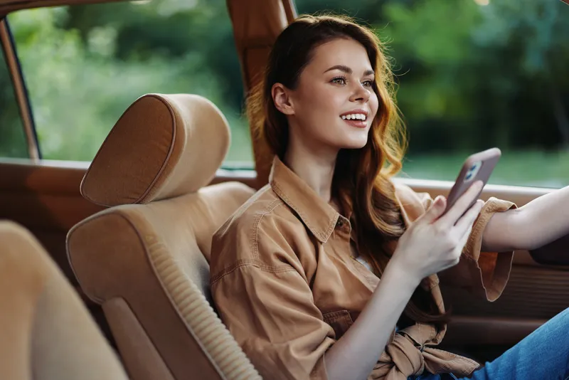 Young woman smiling in a vintage car while using a smartphone, surrounded by green nature, showcasing joy and connection in a relaxed summer atmosphere.