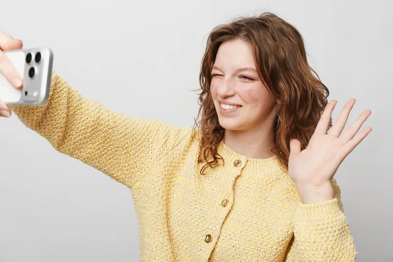 Young woman with curly hair wearing yellow sweater takes selfie with smartphone, smiling and waving hand. Concept of communication, social media, and casual lifestyle.