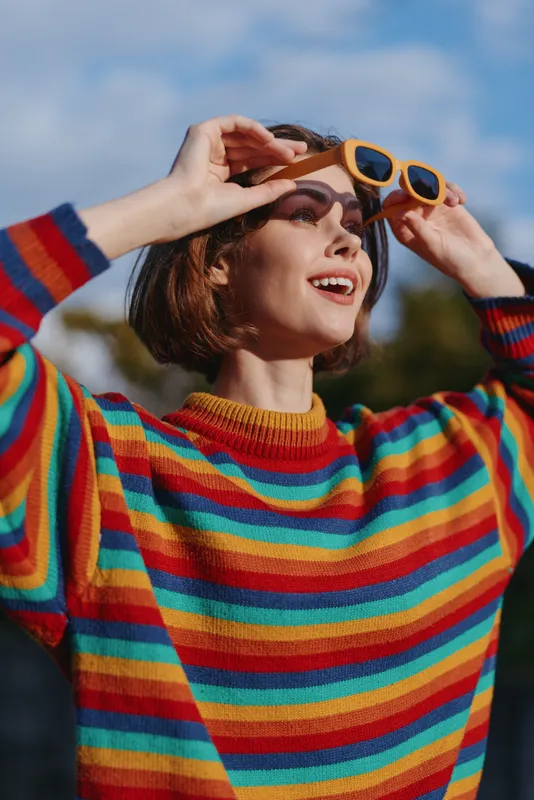 Woman in a colorful striped sweater wearing yellow sunglasses, smile in portrait outdoors while looking up to blue sky, casual fashion, short brown hair and warm sunlight.