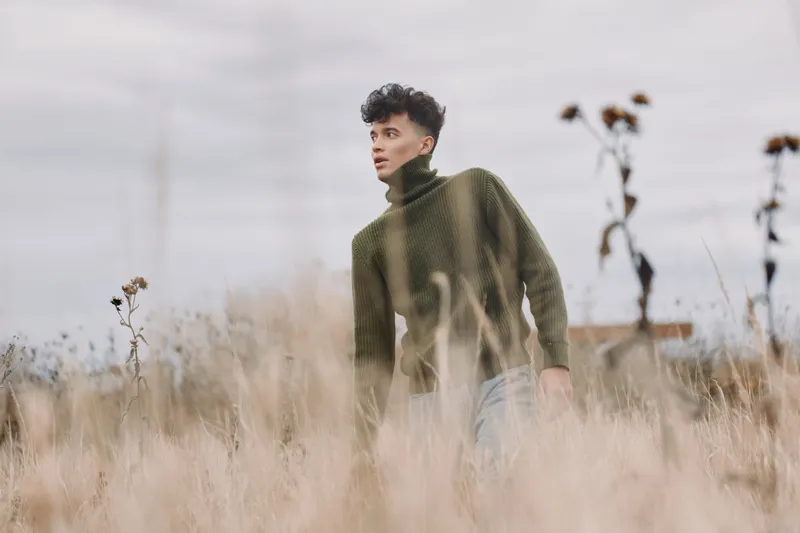 A candid portrait of a young man walking through tall dry grass in a green sweater, expressing quiet introspection and natural authenticity against a soft, overcast sky and muted tones.