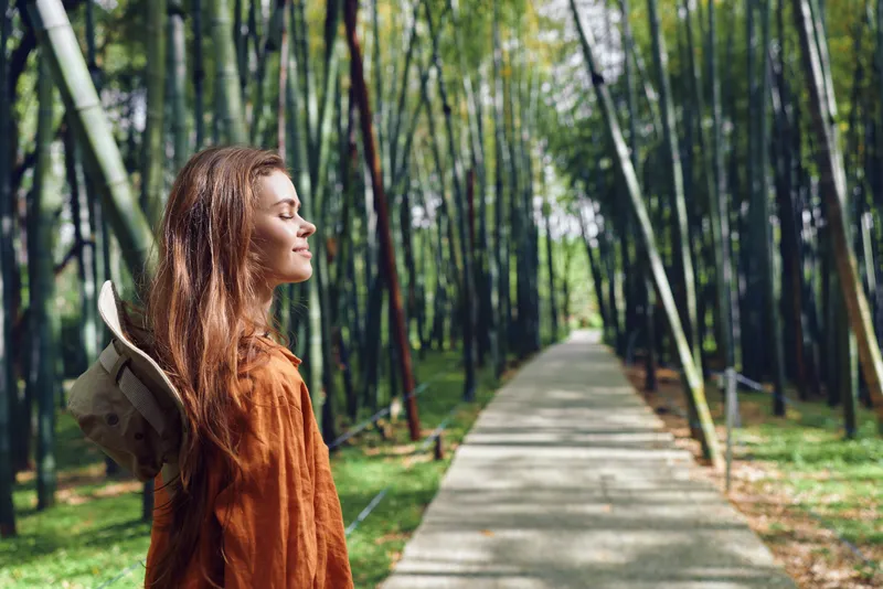 Woman with backpack in bamboo forest path, portrait of peaceful traveler enjoying sunlight and fresh air, eyes closed and smiling, nature walk and serene outdoor moment.