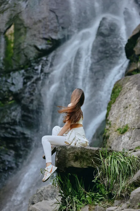 woman sitting by waterfall, flowing water, natural landscape, peaceful, long hair, casual white outfit, outdoor relaxation