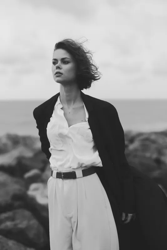 Woman in blazer and white shirt stands on a rugged coast, wind blown hair and bold fashion against a moody seascape
