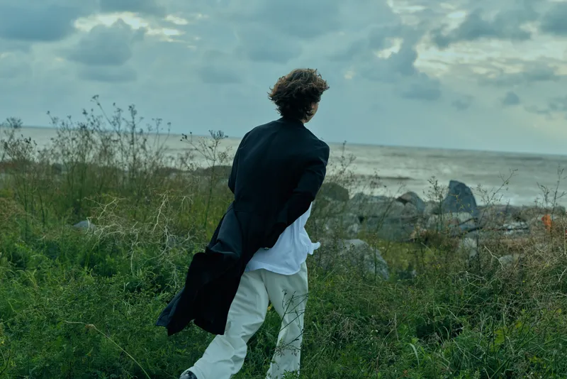 Back view of a man walking along a rugged coastline, jacket fluttering, toward a vast sea and horizon, capturing outdoor nature, wind, and a contemplative, serene travel mood