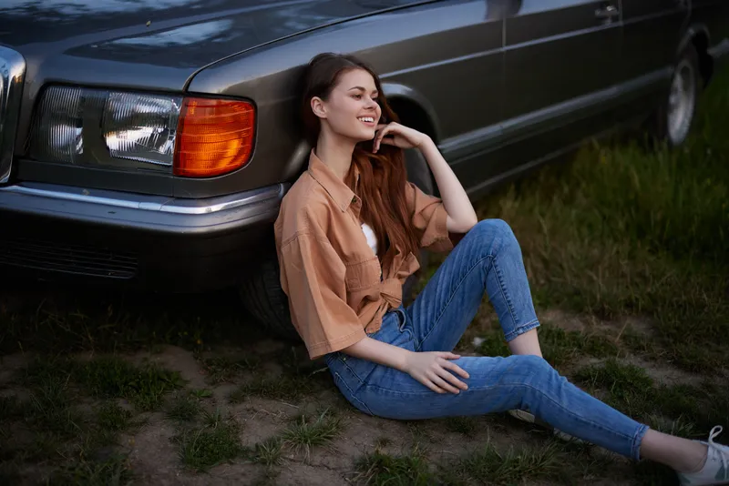 Stylish young woman in casual outfit sitting by vintage car, showcasing relaxed lifestyle, happiness, and summer vibes in a green outdoor setting.