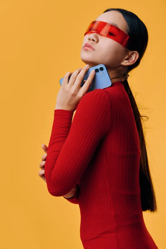 Woman in red sweater with long hair wearing red futuristic sunglasses holding smartphone close to chest against yellow background in studio for technology and fashion.