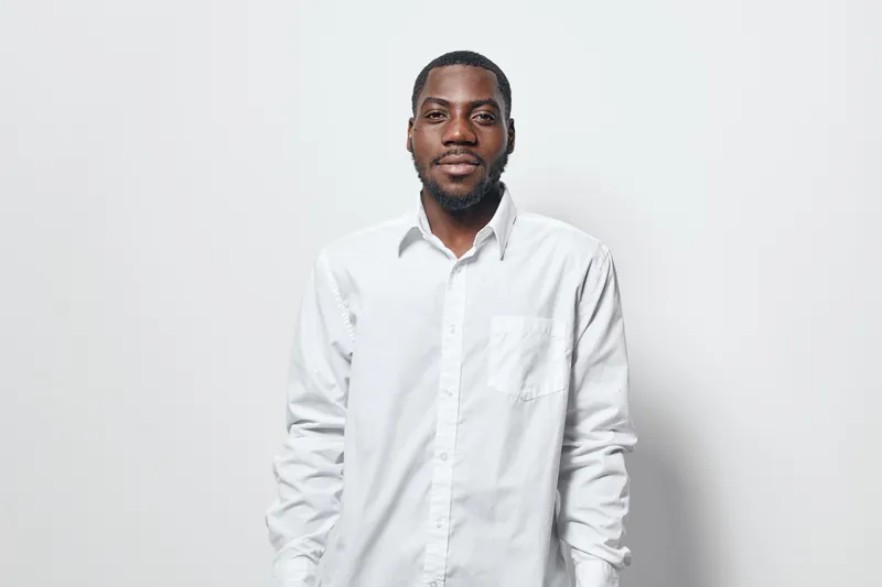African American man with dark skin wearing white shirt standing against white background looking at camera with calm expression and relaxed posture in studio portrait.
