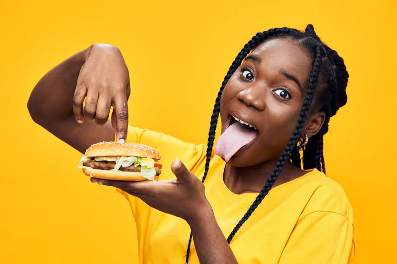 Happy young Black woman enjoying a delicious hamburger with vibrant yellow background, showcasing excitement and love for fast food