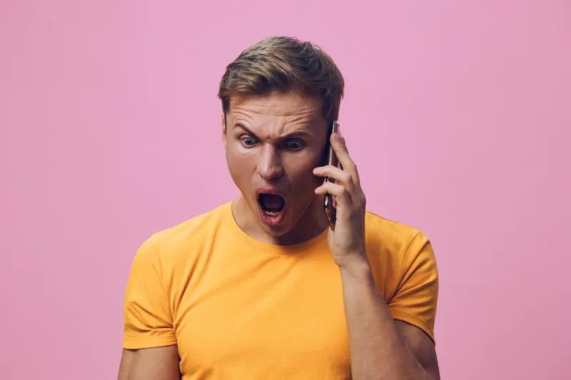 shocked young man in orange tshirt talking smartphone isolated pink background facial expression surprise digital communication concept