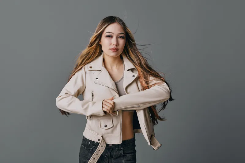 Young woman posing and wearing a beige jacket and black jeans with long hair and neutral expression on gray background in studio setting. Female model with casual style.