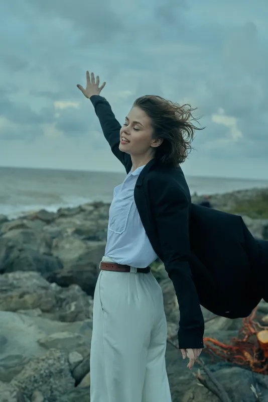 Business woman on a rocky coast raises her arm toward the wind, blazer and white shirt billowing, outdoors scene with ocean horizon, confident movement, fashion inspired professional vibe