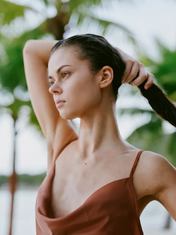 A young woman stands outdoors in warm light, adjusting her hair with a calm, focused gaze. She wears a rust brown top, while palm trees blur softly in a tropical background.
