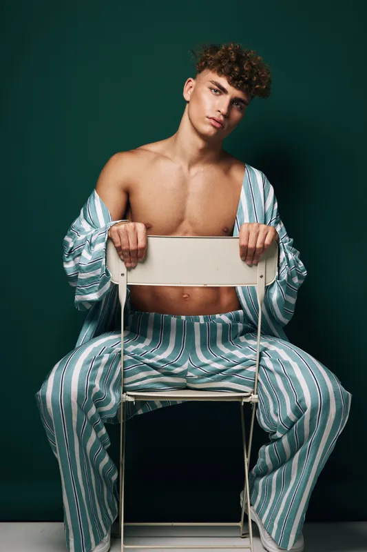 Young man with curly hair and striped pajamas sitting with chair turned backwards against dark green background for style fashion pose and relaxed mood.