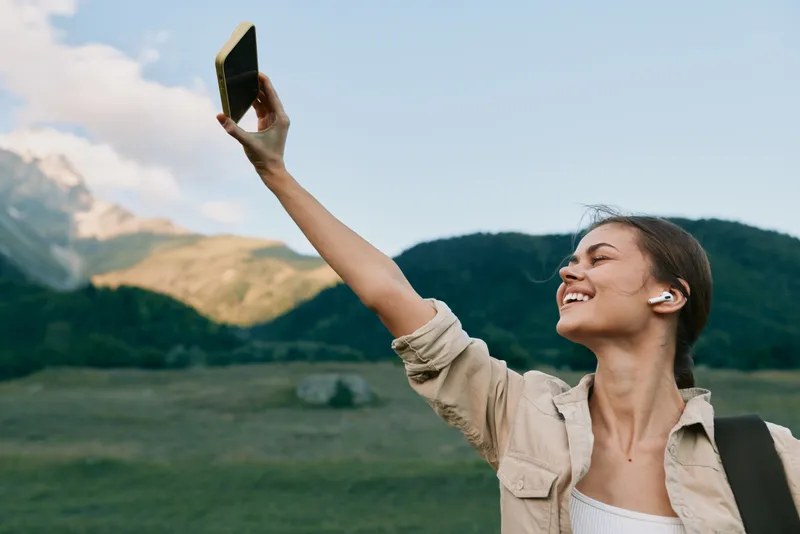 Young woman lifestyle takes selfie with smartphone outdoors in nature mountains background wears casual clothes and wireless earbuds enjoys fresh air and freedom