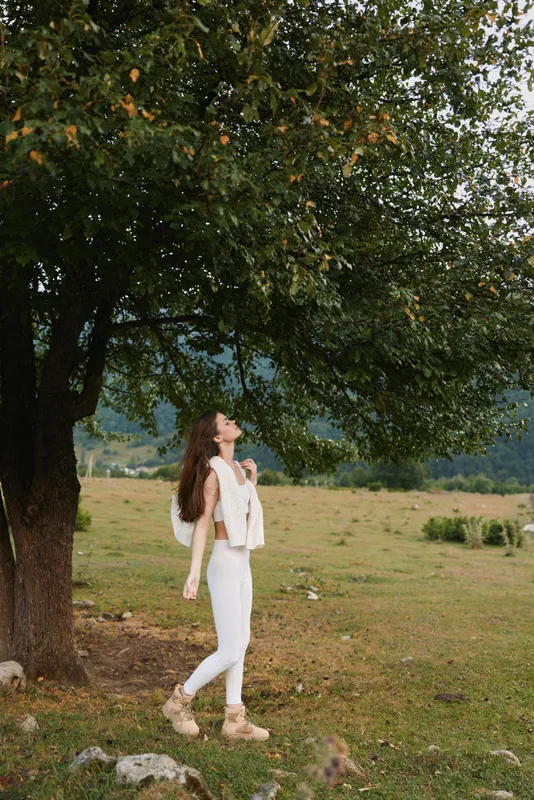 Woman lifestyle portrait outdoors, a figure stands under a large tree in a sunlit field, wearing white outfit, relaxed pose, serene mood and natural setting.