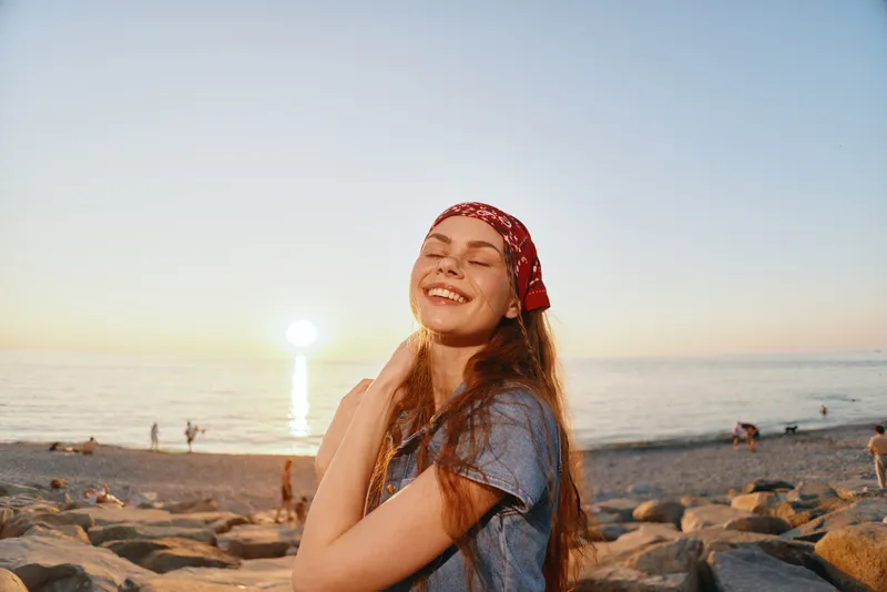 Lifestyle woman enjoying modern boho-western denim street style by the beach at sunset, smiling with relaxed joy and warm film color tones.