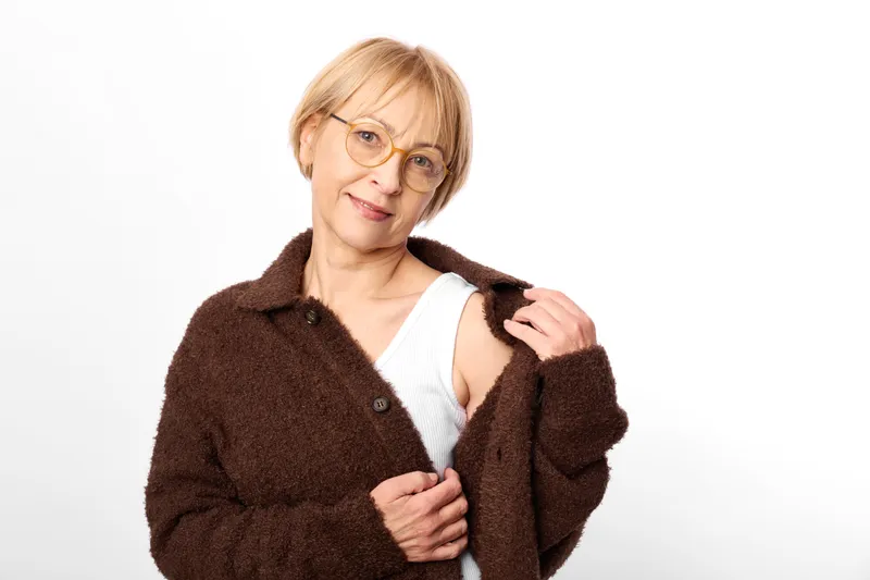 Woman portrait with glasses smiling, wearing brown coat over white top, holding coat lapels, fashion warm style, studio white background, calm confident adult mature female.