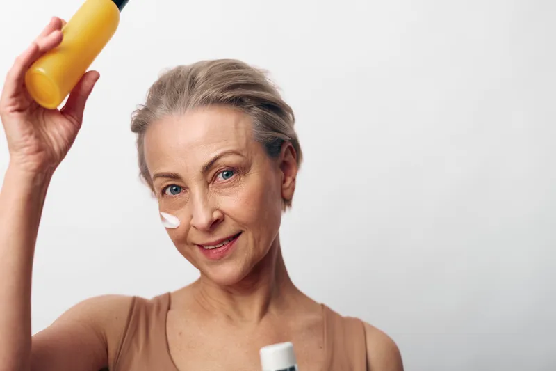 Mature woman with short grey hair applies sunscreen cream on face holding bottle, smiling gently on white background. Concept of skincare, sun protection, aging skin care, health and wellness.