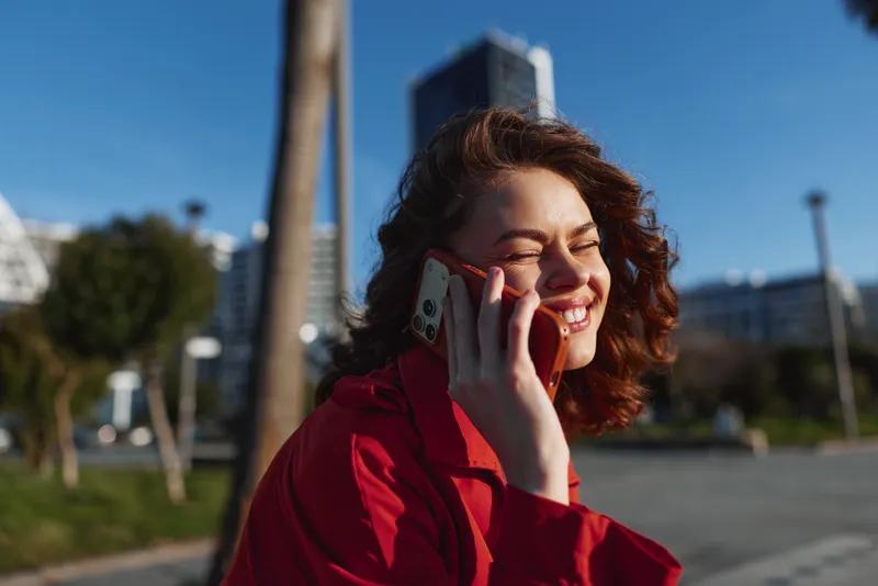 Young woman in a red coat laughs on a sunlit city street while talking on her phone, her bright smile and casual pose framed by blue sky and modern buildings.