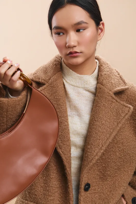 Asian woman wearing a warm coat and knit sweater carries a round tan bag, standing against a neutral beige backdrop to convey a chic, fashion forward mood.